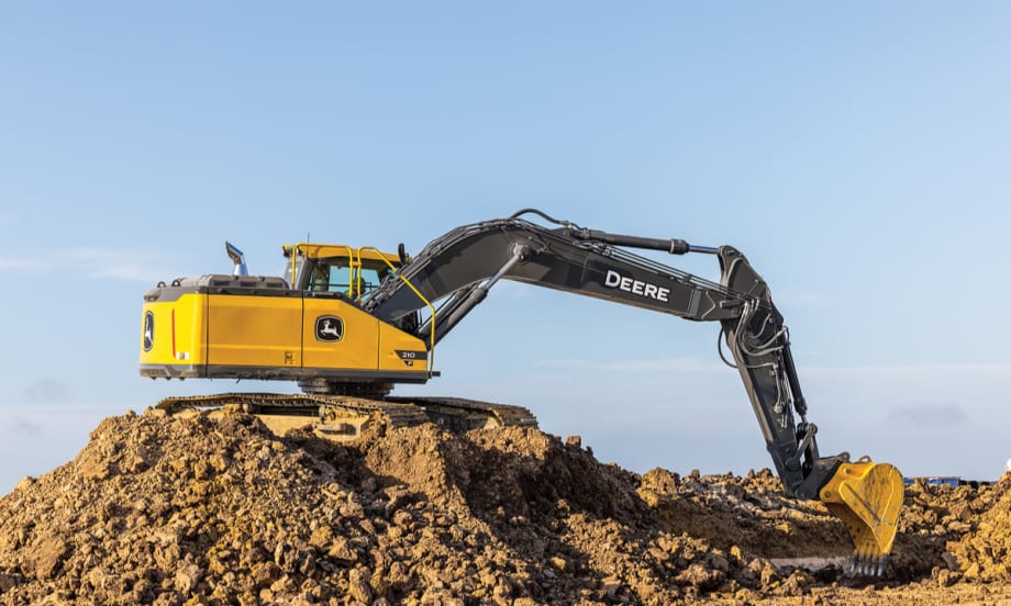 John Deere P-Tier midsize excavator positioned on a dirt mound with the boom and bucket extended at a jobsite.