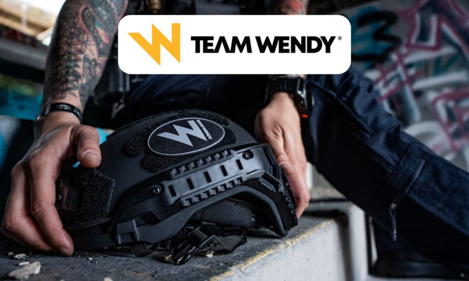 Closeup of hands reaching to pick up a ballistic helmet and a Team Wendy logo in a white box top center in the image.