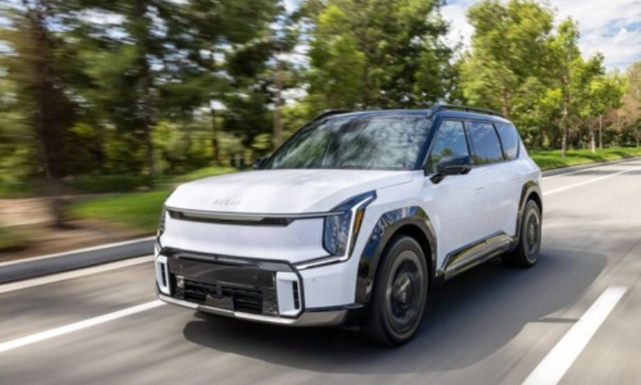 A white 2026 Kia EV9 electric SUV driving on a tree-lined roadway.