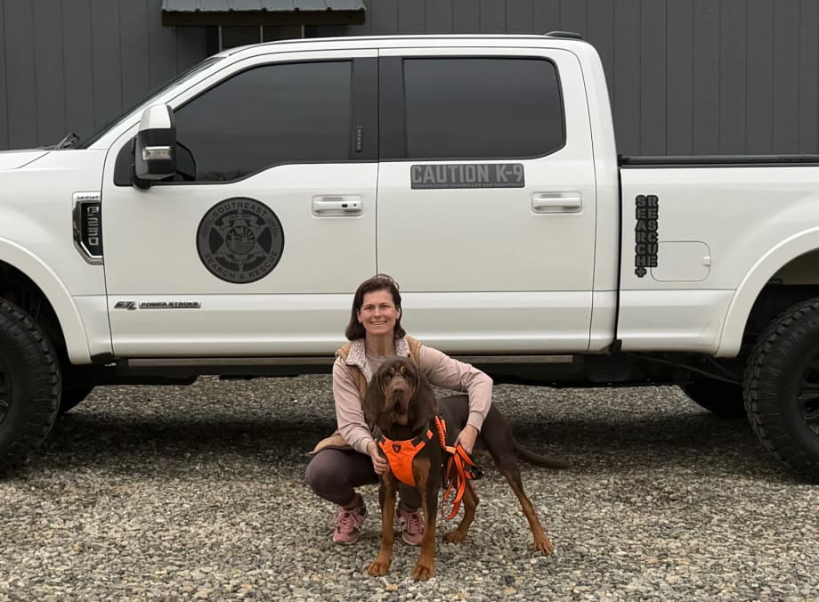 Woman kneeling with a Bloodhound in front of a white pickup truck.