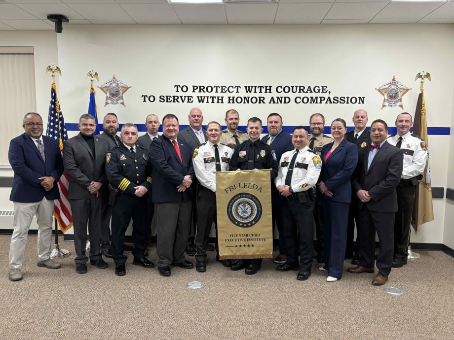 Group of police chiefs in uniform stand in a group at the front of a room following a training class.