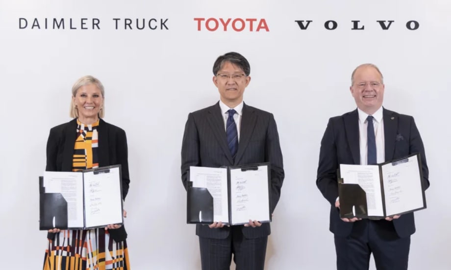 Woman and two men standing holding paperwork agreements they have signed with logos for Daimler Truck, Toyota, and Volvo over their heads.