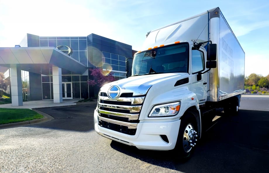 White box delivery truck parked outside a modern office building on a sunny day. The truck is photographed from the front angle, with chrome detailing reflecting sunlight and the building entrance visible in the background.