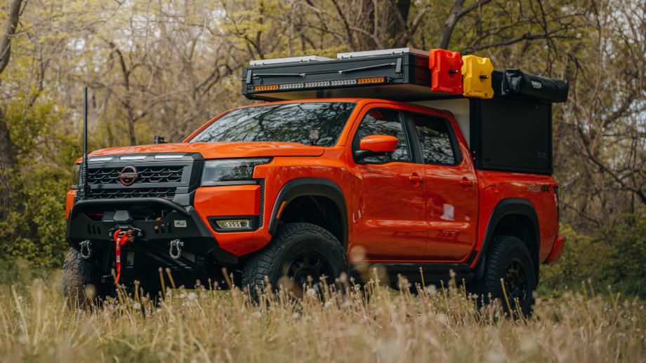 Orange Nissan Frontier PRO-4X overland build parked in a grassy wooded area with off-road tires, heavy-duty front bumper, winch, rooftop cargo storage, and camping accessories mounted above the truck bed.