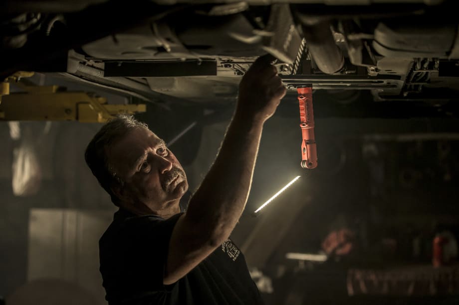 Mechanic working beneath a raised vehicle in a dimly lit garage, illuminated by a hanging Streamlight work light. The technician reaches toward the undercarriage while inspecting or repairing automotive components.
