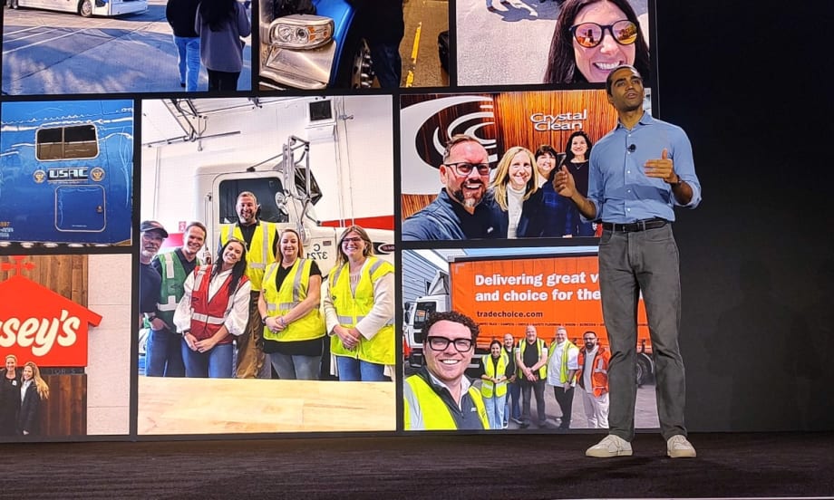 Presenter speaking onstage in front of a large display wall featuring fleet and transportation team photos, including trucks, drivers, and company groups wearing safety vests.