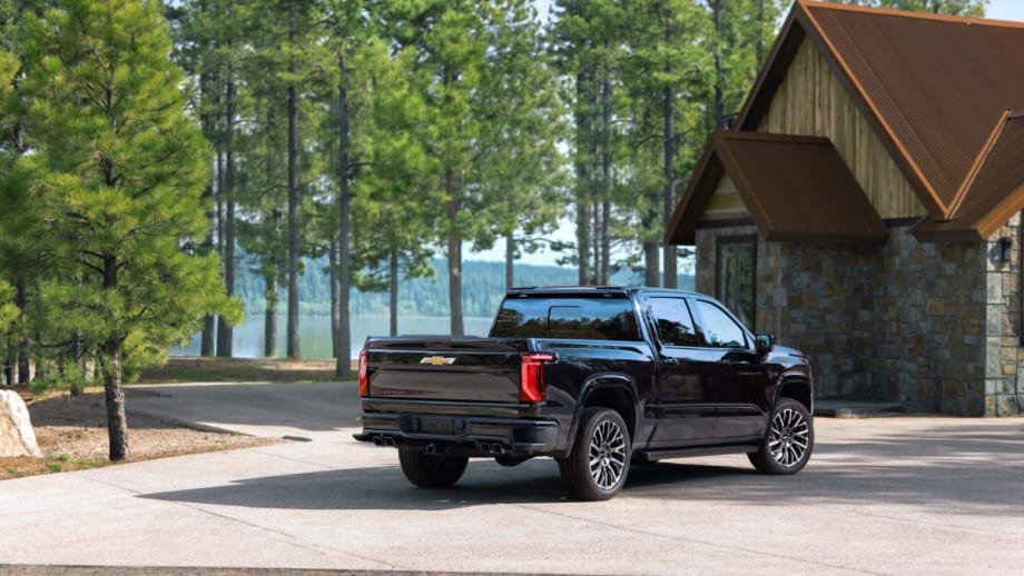 Rear three-quarter view of a black 2027 Chevrolet Silverado 1500 parked outside a lakeside cabin surrounded by pine trees.