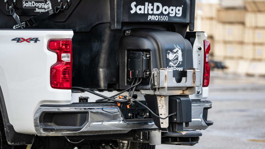 Close-up of a SaltDogg PRO1500 tailgate salt spreader mounted on the rear of a white 4x4 pickup truck, showing the hopper, spreader assembly, wiring connections, and mounting hardware.