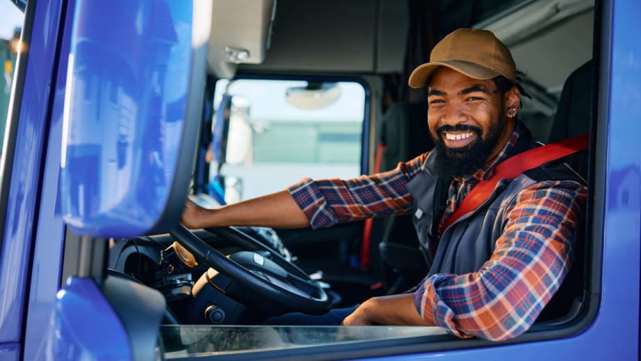 Commercial truck driver wearing a red seat belt smiles from the driver’s seat of a blue truck.