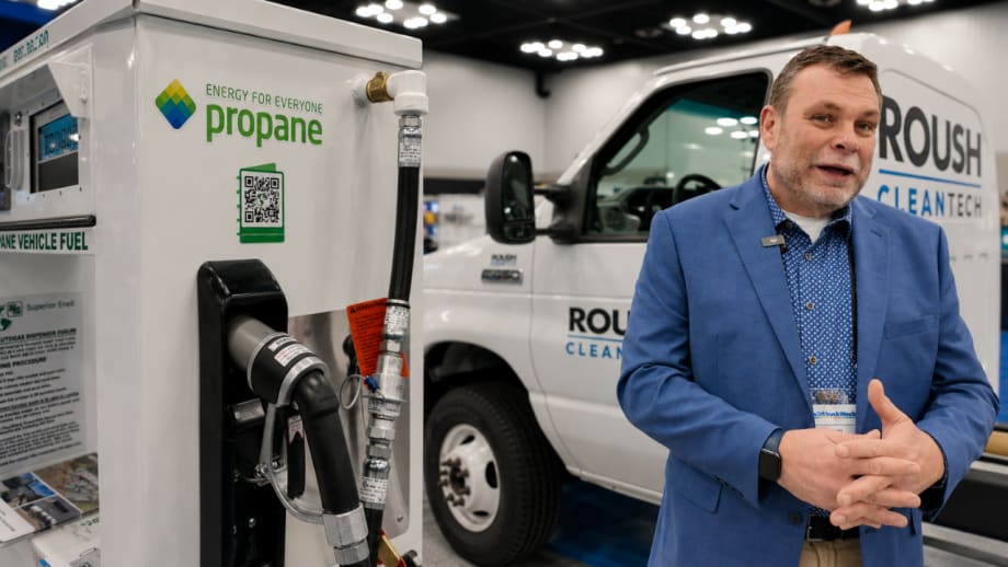 A man in a blue blazer stands beside a ROUSH CleanTech truck and propane autogas fueling dispenser at an industry trade show.