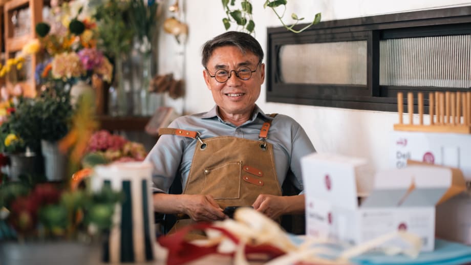 Flower shop owner wearing glasses and an apron sits behind a worktable surrounded by flowers, packaging supplies, and small boxes.