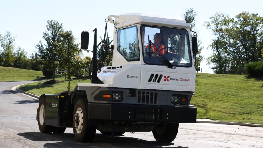Kalmar Ottawa electric terminal tractor drives on a paved roadway with an operator in the cab, showcasing the zero-emission yard truck in an outdoor commercial fleet setting.