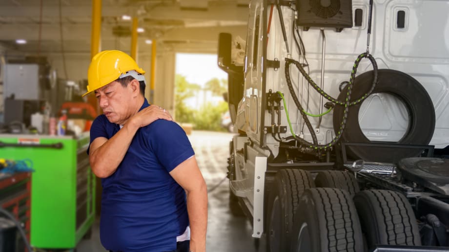 Fleet technician wearing a yellow hard hat holds his shoulder beside a white truck inside a maintenance shop.
