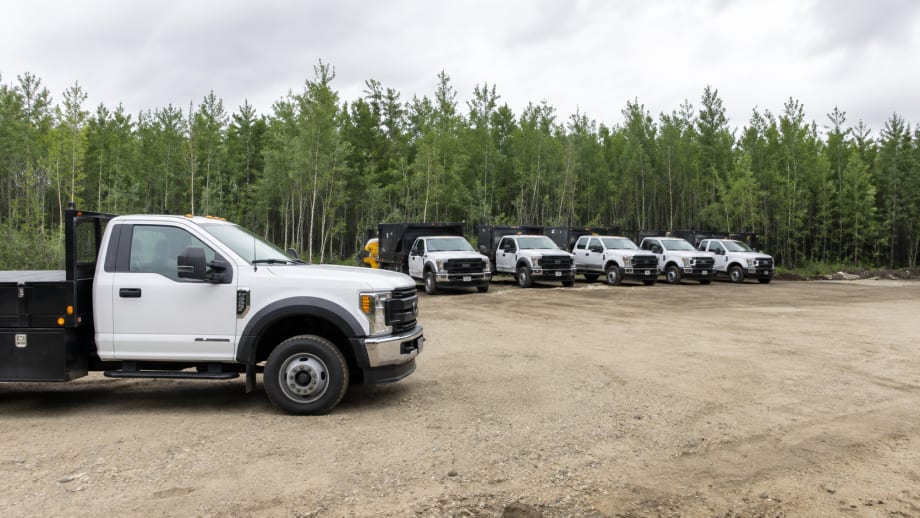 Row of white work trucks with black utility bodies parked on a gravel lot beside a tree line under an overcast sky.