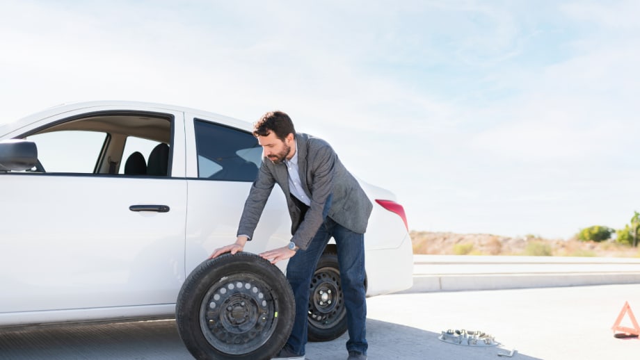 Driver lifts a replacement tire beside a white sedan stopped on the roadside, with tools and a warning triangle nearby.