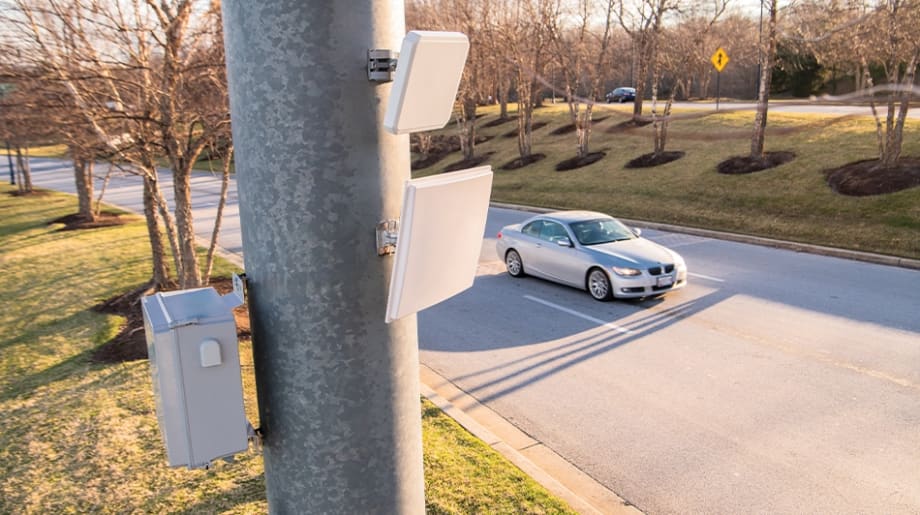 Roadside traffic monitoring equipment mounted on a utility pole overlooks a passing car, illustrating wireless vehicle detection technology used for traffic management, signal optimization, or roadway data collection.