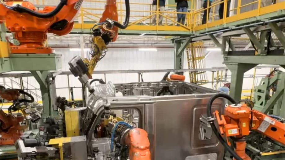 Industrial manufacturing facility with multiple robotic arms assembling a heavy-duty truck cab on an automated production line, while workers observe from an elevated platform above the assembly area.