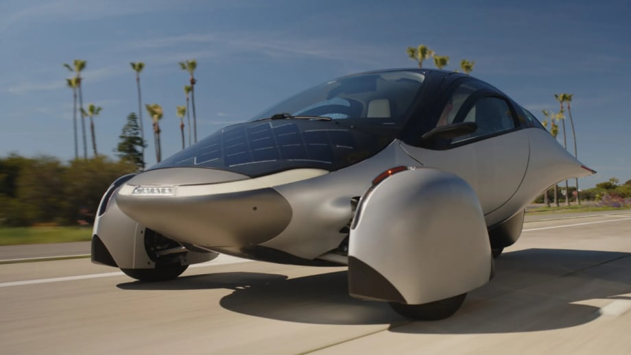 Futuristic silver three-wheeled Aptera solar electric vehicle drives along a palm-lined road, featuring integrated solar panels, aerodynamic bodywork, enclosed front wheels, and a streamlined teardrop-shaped cabin.
