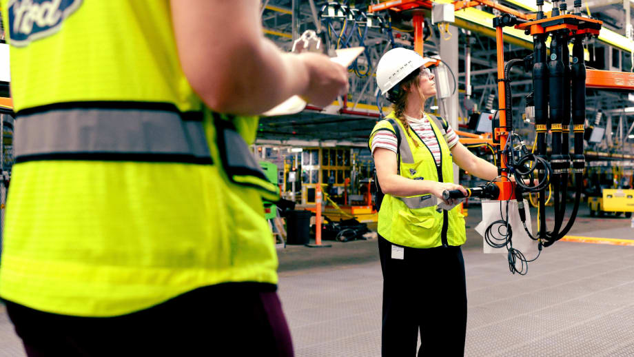 Ford plant employee wearing a safety vest and hard hat operates production equipment while another worker stands nearby with a clipboard.