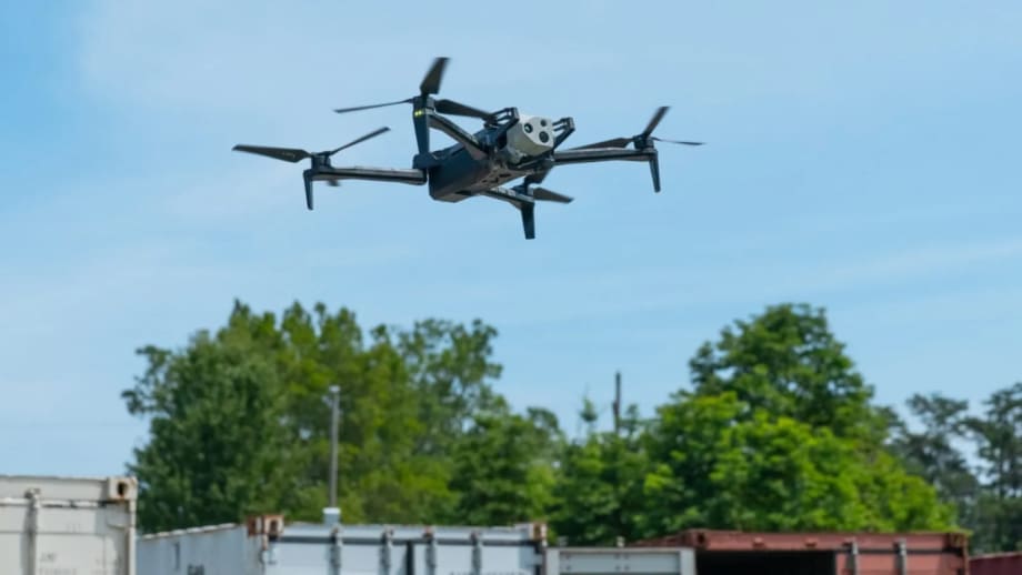 A HAWK drone flies above an industrial area with trees, buildings, and shipping containers visible below.