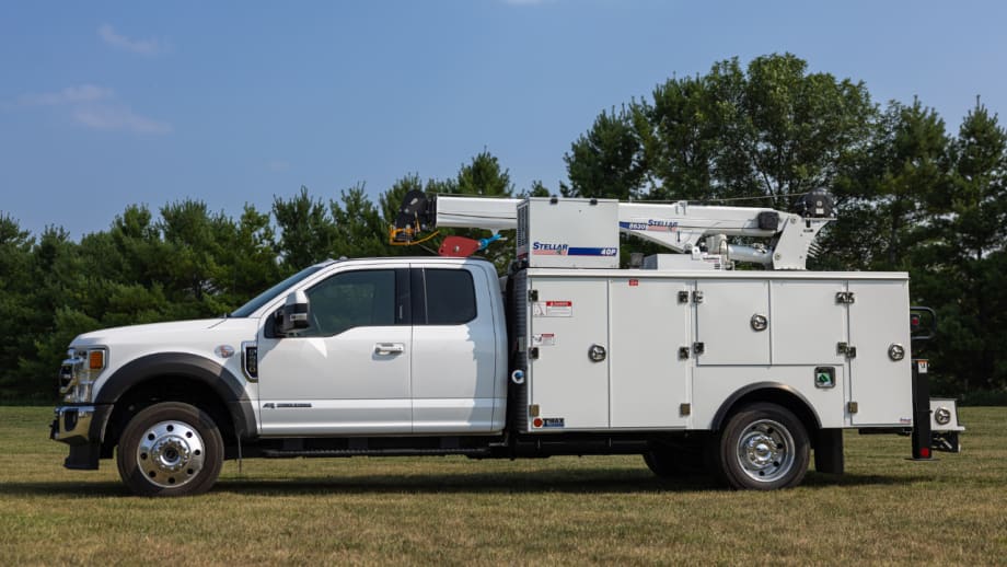 White utility service truck equipped with a Stellar crane and service body, parked on grass with trees in the background.