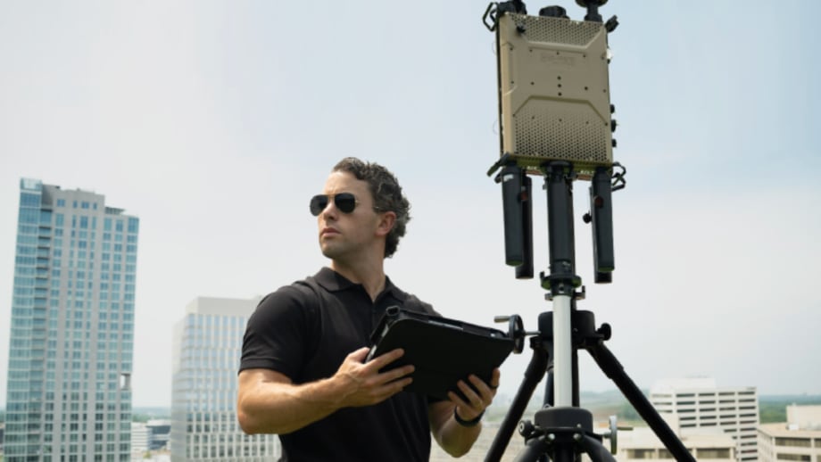 A technician wearing sunglasses holds a tablet beside tripod-mounted electronic detection equipment on a rooftop, with office buildings visible in the background.
