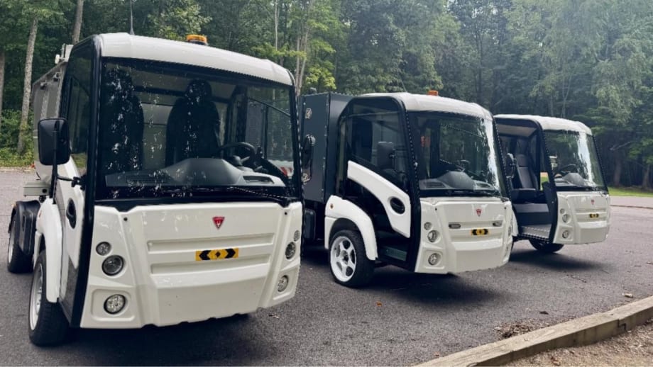 Three compact white utility trucks parked in a row on a paved lot, surrounded by trees, with large windshields, enclosed cabs, and amber warning lights.