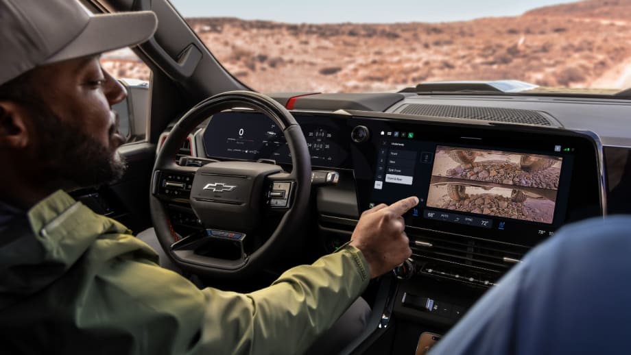 Driver inside a Chevrolet Silverado using the center touchscreen to view underbody camera images while parked on rocky terrain.