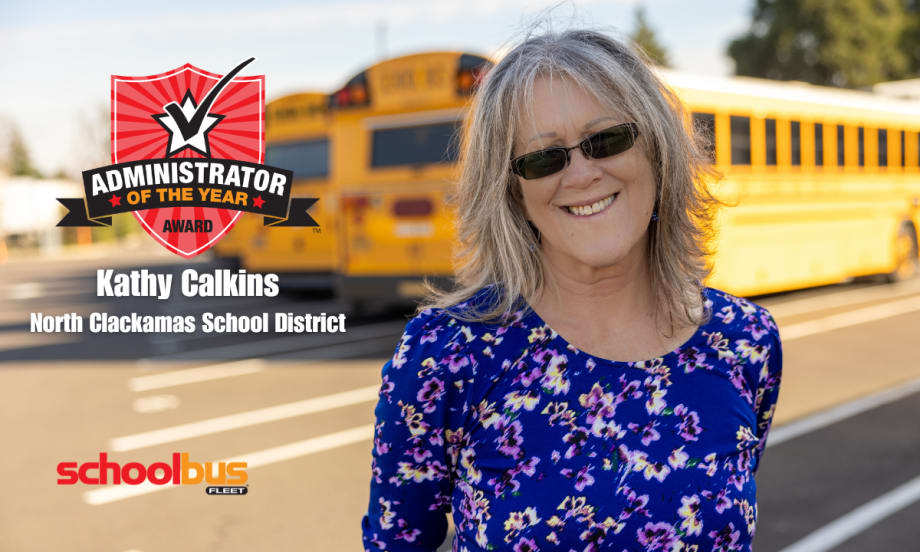 Kathy Calkins stands in a parking lot with school buses behind her and the admin of the year award logo next to her