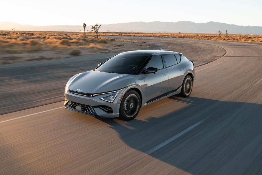 Gray 2027 BMW iX3 electric SUV driving on a desert highway with mountains and Joshua trees in the background at sunset.