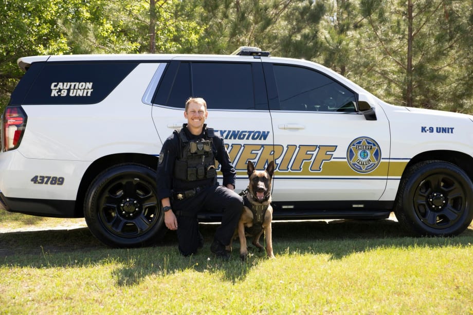 K-9 police officer with German Shepherd beside sheriff's white patrol vehicle