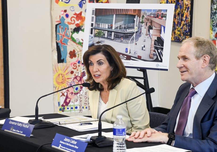 Governor Kathy Hochul sitting at a table with a microphone, Janno Lieber sits to her left.