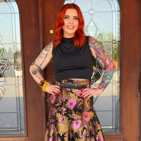 A woman with red hair and her arms akimbo, standing in front of glass doors.