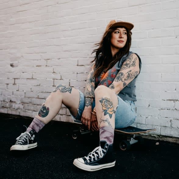 A young woman in jean shorts, wearing a baseball hat, leans against a brick wall.