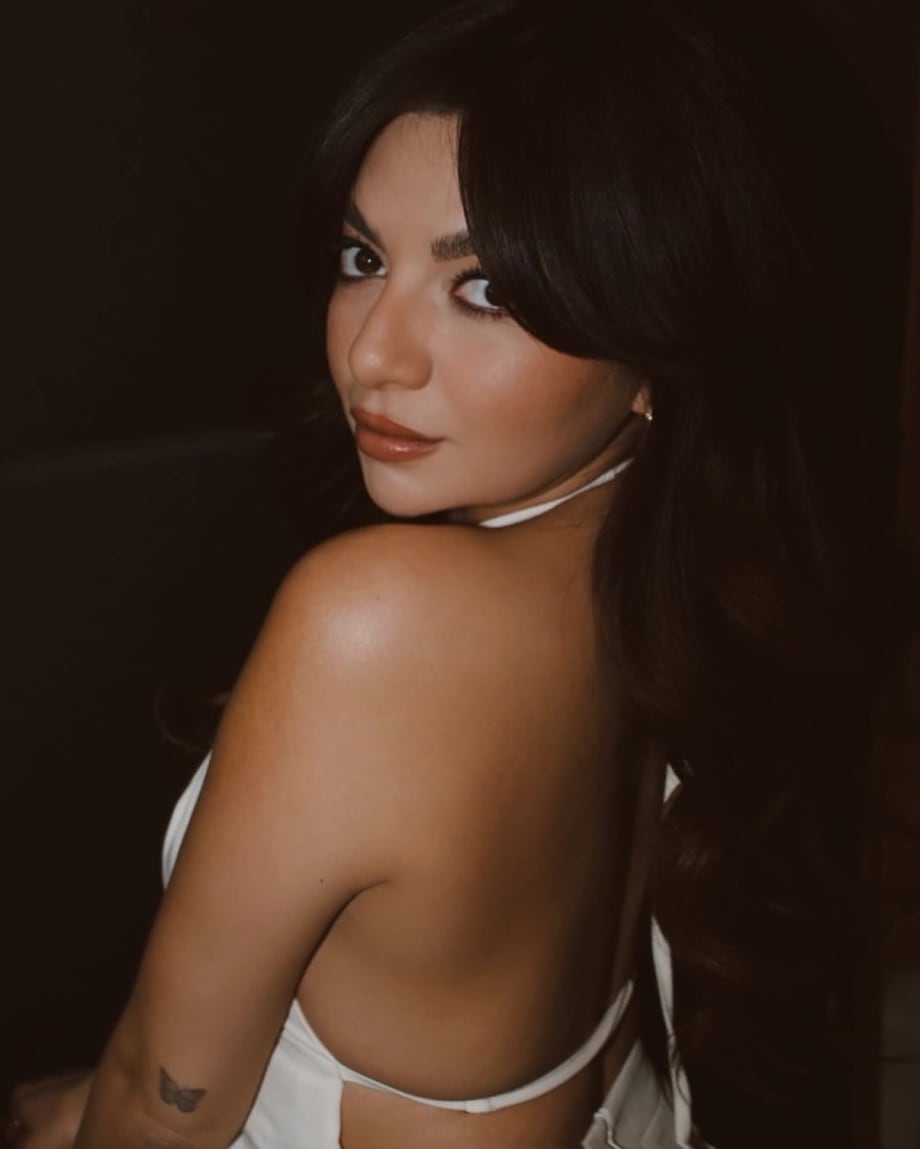 A young woman with long brunette hair looks over her shoulder while wearing a white halter top.
