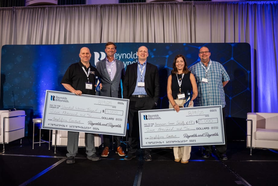 Group of five people on stage holding large checks at a Reynolds and Reynolds event, recognizing winners of an automotive innovation contest.