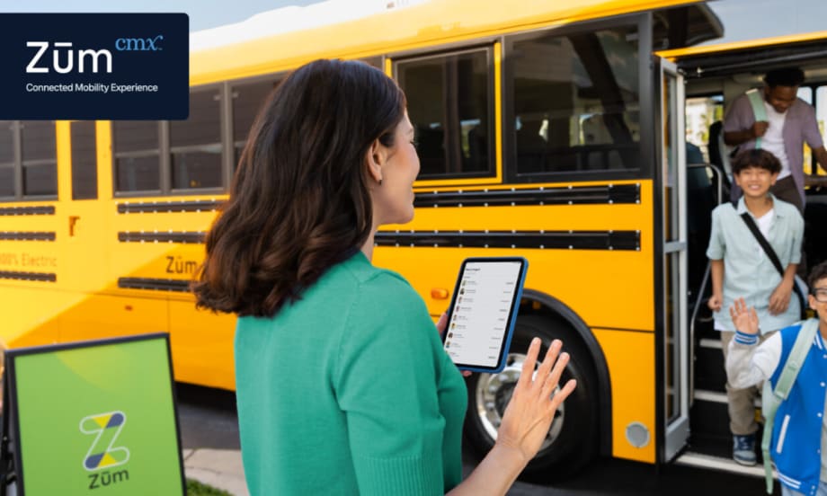 A woman holds a tablet and waves at children disembarking a school bus.