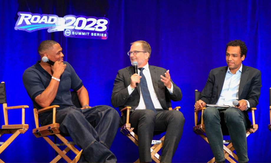 Three speakers participate in a panel discussion at the Road to 2028 Summit Series. A man in a gray suit speaks into a microphone while two other panelists listen on stage beneath a "Road to 2028 Summit Series" sign.