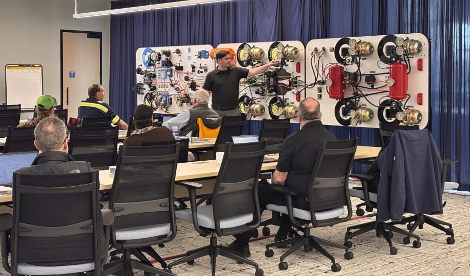 Instructor leads a brake system training class with technicians seated at tables, using a large demonstration board with air brake components and wiring.