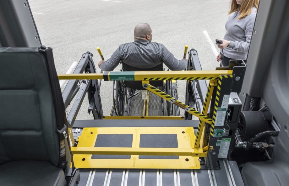 A wheelchair user operates a school bus wheelchair lift equipped with BraunAbility’s LiftSentinel barrier system as another person stands nearby with a handheld control.