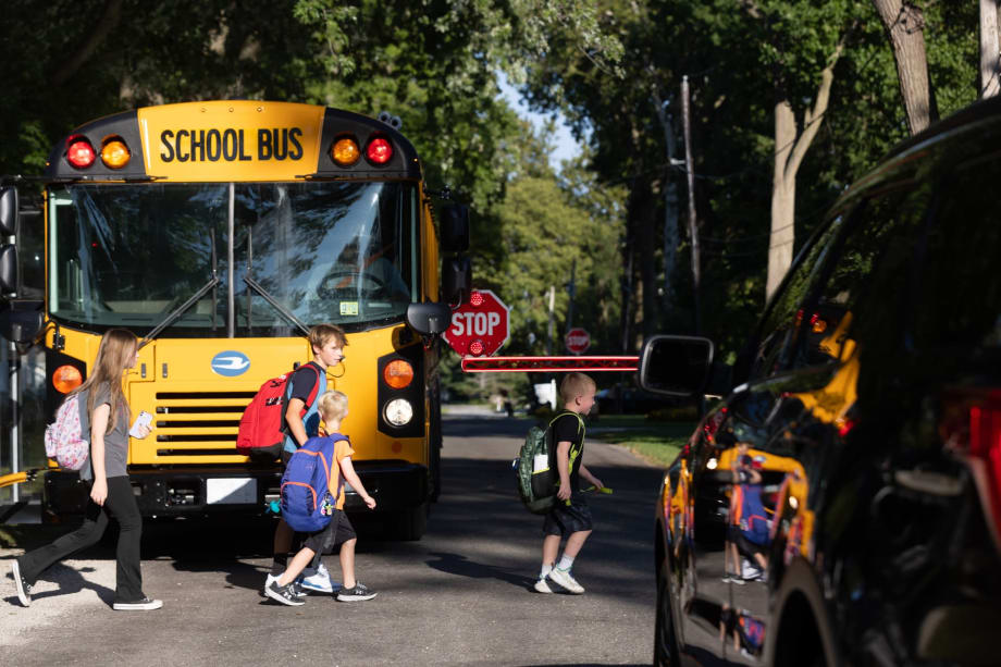 Children cross in front of a stopped school bus with its stop arm extended while a nearby vehicle waits, illustrating school zone safety and risks of illegal passing.