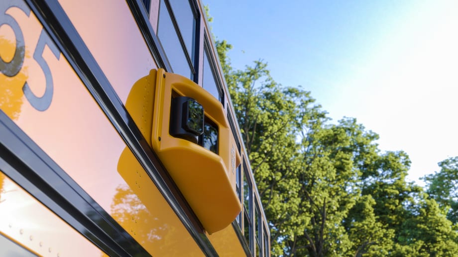 Close-up of a school bus stop-arm camera mounted on the side of a yellow bus, used to record drivers who illegally pass while students board or exit.