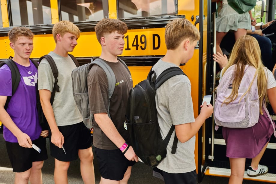 Students wearing backpacks line up to board a yellow school bus, each holding an NFC-enabled student ID card to scan as they enter. The image illustrates student ridership tracking technology that records boarding activity in real time.