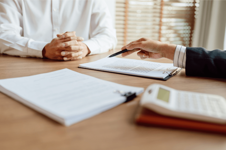 Two people sit across from each other at a desk during a business meeting. One person, wearing a white shirt, has their hands folded while the other gestures with a pen toward documents clipped to a clipboard. Additional paperwork and a calculator are visible on the table, suggesting a discussion involving contracts, finances, or administrative paperwork. Sunlight filters through window blinds in the background, creating a professional office setting.