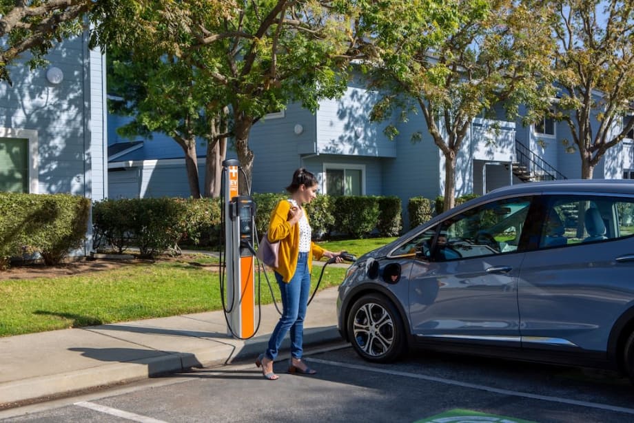 Woman plugs an electric vehicle into a ChargePoint charger outside an apartment complex, representing expanded EV charging access for multifamily housing.