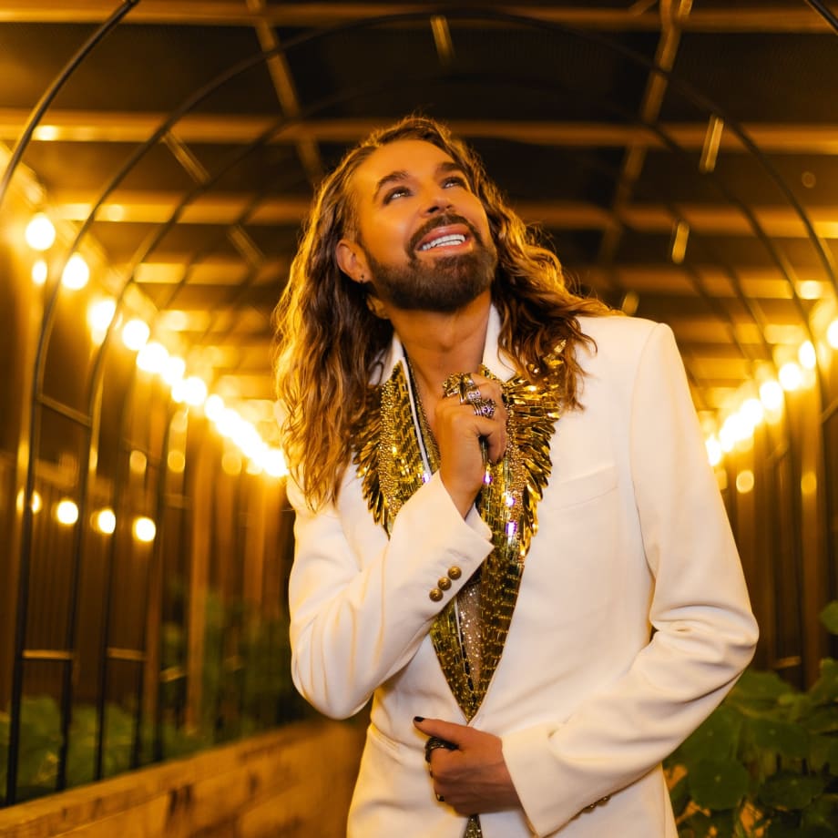A man with long blonde hair, wearing a white tuxedo jacket, looks upwards.