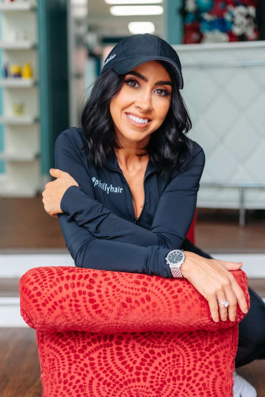 A woman wearing a black cap, leans on a red chair and smiles with folded arms.