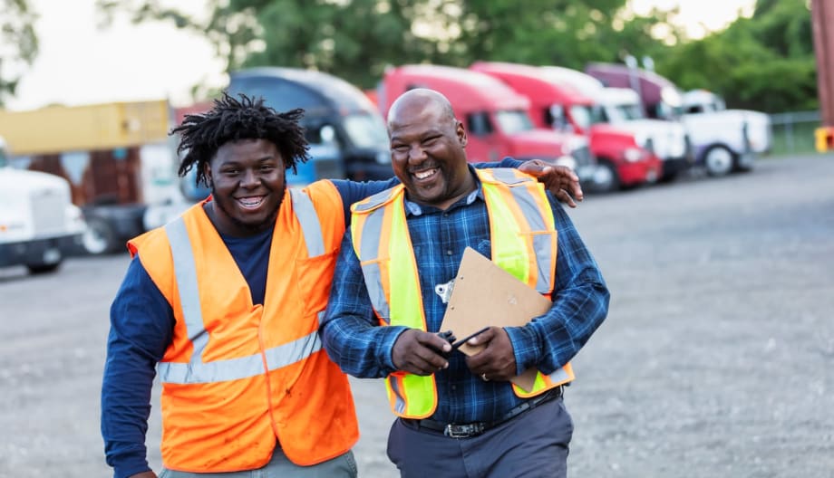 Two black men in safety vests walking together laughing in a truck fleet yard