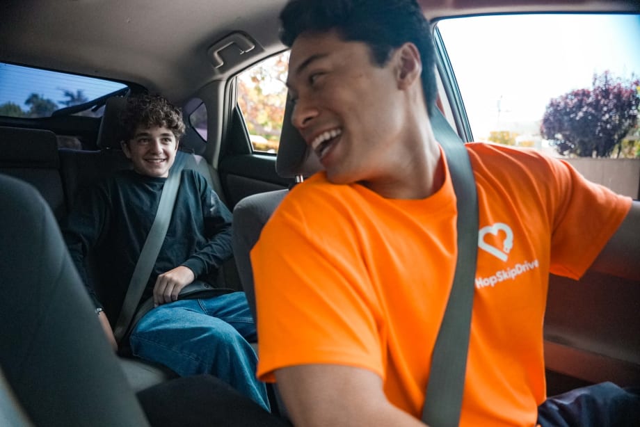 HopSkipDrive driver in an orange shirt smiles at a seat-belted student riding in the back seat of a car.