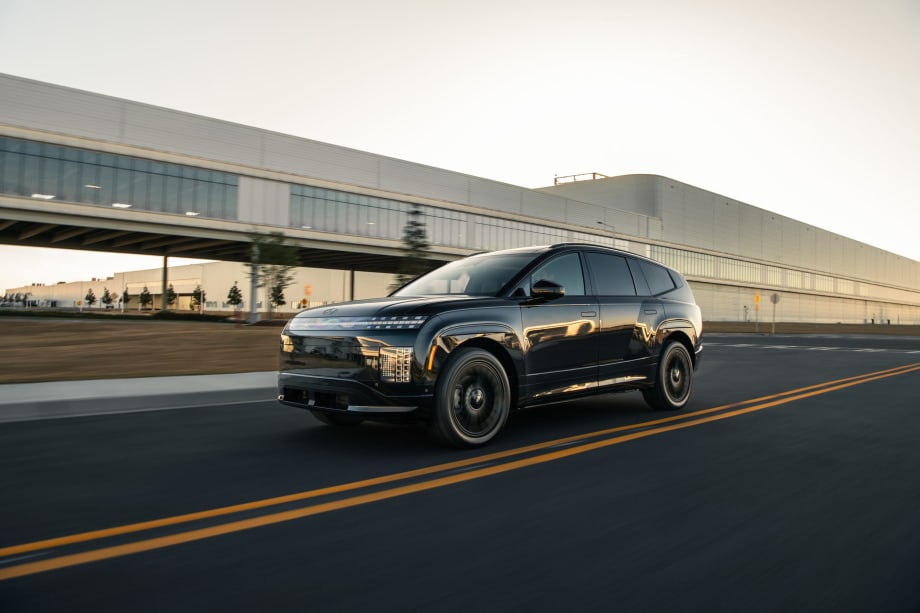 Black Hyundai IONIQ 9 electric SUV driving on a road near a modern industrial building.